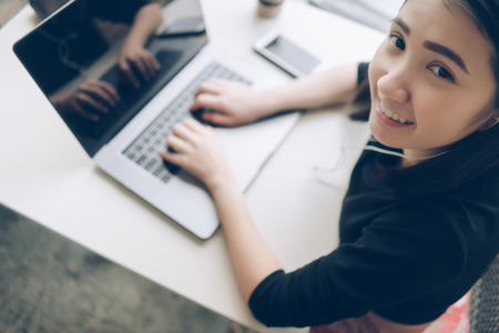 Young business woman smiling using laptop on wooden table while sitting in coffee shop, top view. Happy cute Asian girl using notebook computer looking at camera, listening music on earbuds, media entertainment relaxation concept.の写真素材