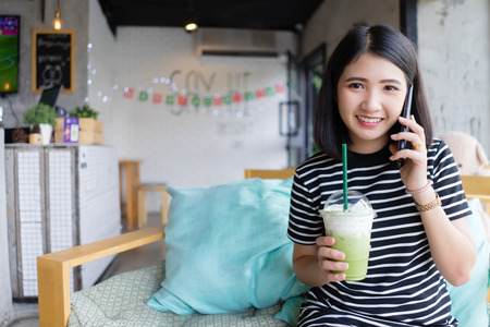 Attractive Young Woman Talking on Mobile Phone at coffee shop  drinking green tea while sitting on the sofa with Happy Facial Expression.
Asian girl relaxing on couch in cafe. Technology, communication relaxation conceptの写真素材