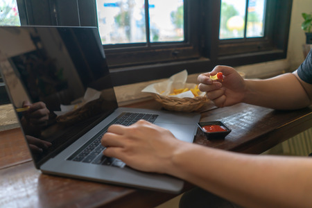 Business man using laptop computer, hand typing notebook keyboard and eating fast food French fries with ketchup.
Sedentary working lifestyle fat with unhealthy eating junk food habits concept.の写真素材
