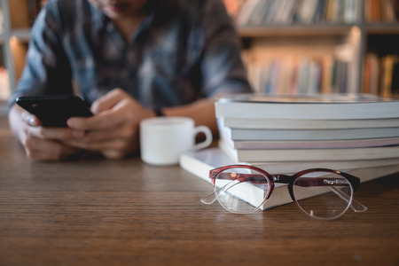 Close up eyeglasses and books stack on wooden desk in university or public library with man using...