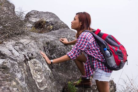 Young climber woman hiking mountain. Asian girl hiker climbing on rock mountain peak cliff.Female hiker with backpack climbs steep rocky hill.の写真素材