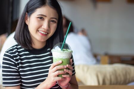 Smiling woman holding matcha green tea latte at coffee shop. Asian girl holding green tea glass in cafe morning. Focus on green tea glass.の写真素材