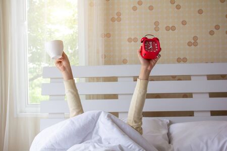 Woman arm hands holding coffee cup and blue alarm clock in on bed in bed room. Young girl with two hands holding blue clock and cup, drinking coffee after awaking. wake up with fun in morning concept.の写真素材