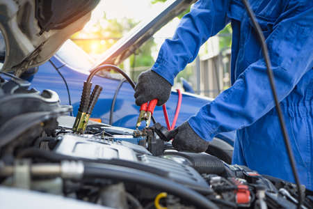 Battery car dead concept. A mechanic repair car battery, charging with red and black cables. fixing vehicle engine power down. Man holding jumper cable in garage.の写真素材