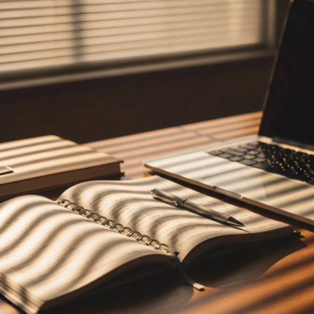A close-up shot of an open notebook and a laptop on a wooden desk, with sunlight casting striped shadows from the blinds.の素材