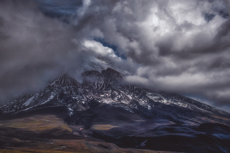 Picturesque mountain landscape with steep mountain peak covered with snow under dense dark clouds