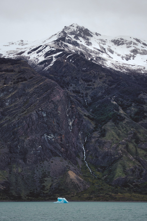 Small blue iceberg floating on water, viewed from the distance against steep mountain, covered with snowの写真素材
