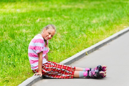A smiling teen girl in roller skates sitting on the asphalt path.