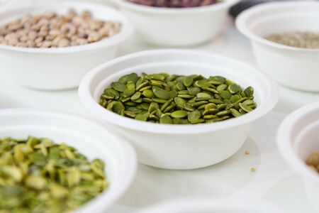 Peeled pumpkin seeds lie in a white plastic plate. Around in the same plates are various seeds, nuts and legumes.の写真素材