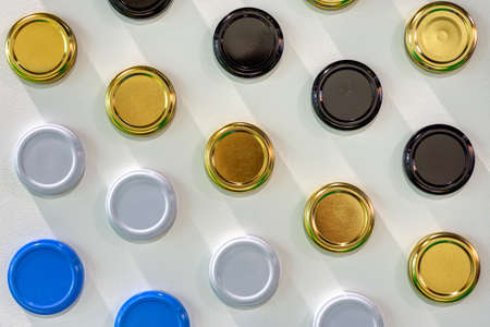 Multi-colored metal lids for glass jars. Covers lie in even rows on a white surface.