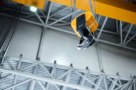 Huge hook assembly of bright yellow moving crane hangs under ceiling in workshop of production plant...