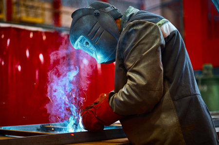Skilled worker in protective mask welds metal part at plantの写真素材