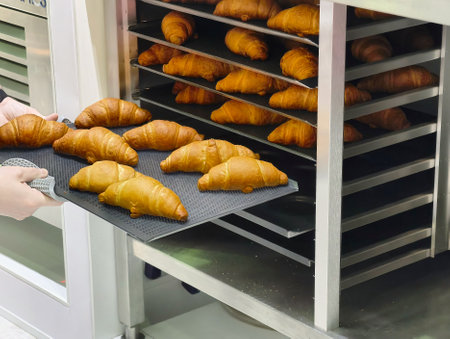 Freshly baked croissants being taken out of the oven in a bustling bakery, showcasing golden layers and irresistible aromaの写真素材