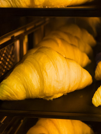 Golden croissants rise in the warm glow of the commercial oven during a busy morning baking sessionの写真素材