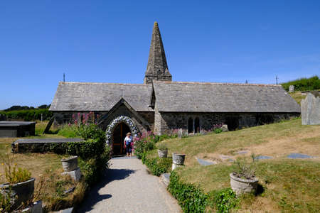 21 Jun 2018. St Enodoc's Church, Trebetherick, Cornwall, UK, under bright and clear blue sky. The footpath leads to the floral adorned entrance. A lady dressed in a white shirt, waits at the doorway.のeditorial素材