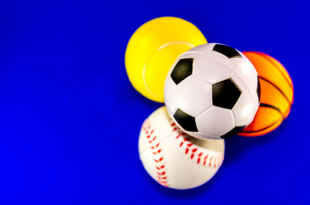 A closeup image of a stack of sports balls against a reflective blue background. In the stack is a football, baseball, basketball and tennis ball.の写真素材