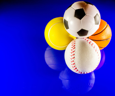 A closeup image of a stack of sports balls against a reflective blue background. In the stack is a football, baseball, basketball and tennis ball.の写真素材