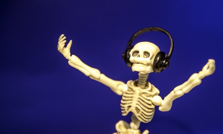 A skeleton enjoys listening to his / her music on their headphones. Taken on a blue background.