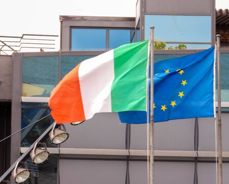 Both the Italian and European flags flying together, with buildings behind.の写真素材