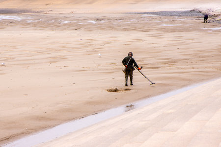 An adult male walking along the beach with a metal detector to seek our treasures hidden below the...