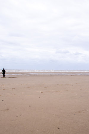 An adult male walking along the beach with a metal detector to seek our treasures hidden below the...