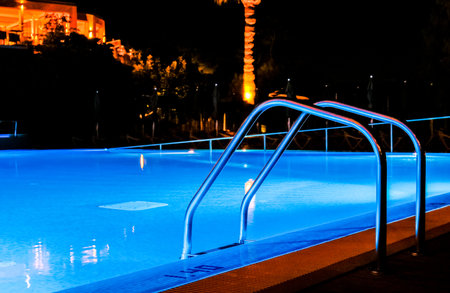 A resort swimming pool set of stairs that allow access in and out of the pool, seen at night lit by the pool lights.の写真素材