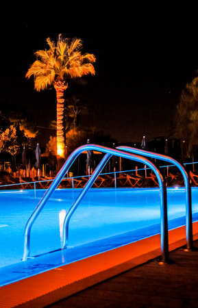 A resort swimming pool set of stairs that allow access in and out of the pool, seen at night lit by the pool lights.の写真素材