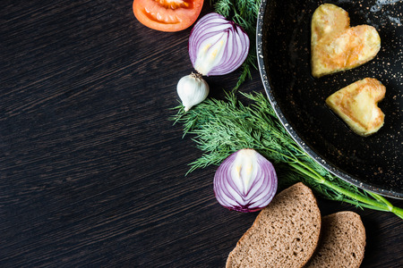 Fried eggs in the shape of a heart in a frying pan, near which lay bread slices, red onion, tomato, garlic and dill on dark wooden tableの写真素材