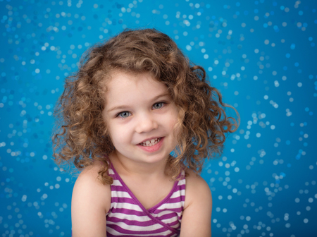 Happy smiling laughing child looking at camera: girl with curly hair against blue background with...