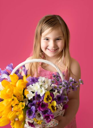 Smiling girl standing with a basket of spring flowers looking at the camera, pink backgroundの写真素材