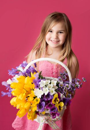 Smiling girl standing with a basket of spring flowers looking at the camera, pink backgroundの写真素材