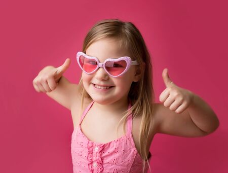 Happy smiling girl in heart shaped pink glasses giving thumbs up; success or achievementの写真素材