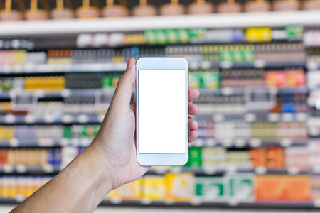 Man's hand shows mobile smartphone with white screen in vertical position, Supermarket blurred background - mockup template and clipping pathの写真素材