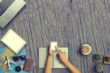 Concept of hipster travel traveler travelling and shopping. Hand Using Smartphone with  Cup of Coffee, cactus, laptop computer and passport on wooden table background.Copy Space and Vintage toneの写真素材