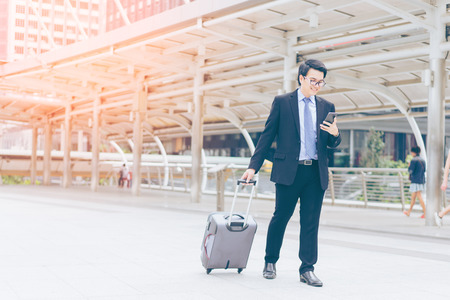 Young asian handsome businessman and glasses using smartphone while walking outside business district before work travel with suitcase and smiling at business districtの写真素材