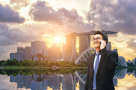Portrait of asian handsome business man using cell phone, smiling, Singapore downtown business district background.の写真素材