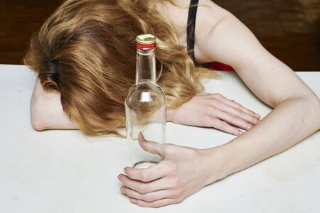 Drunk woman holding an bottle vodka and sleeping with her head on the tableの写真素材