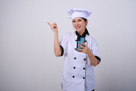 Photo of excited happy female Asian chef using mobile phone pointing on empty space or copy space for product isolated on white backgroundの写真素材