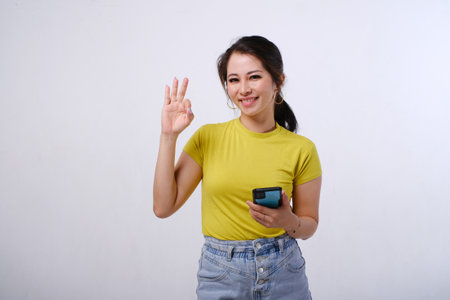 Photo of smiling female Asian using mobile phone showing OK hand sign gesture of approval on isolated white backgroundの写真素材
