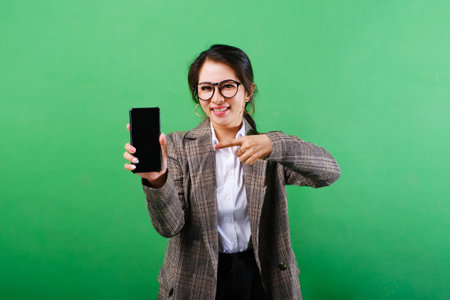 Photo of smiling Asian businesswoman showing mobile phone screen with hands pointing on blank phone screen on isolated green backgroundの写真素材
