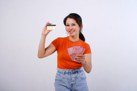 Smiling Asian woman wearing casualÂ shirt holding credit card and Indonesian money isolated on white backgroundの写真素材