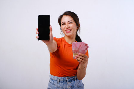 Excited and happy Asian woman wearing casualÂ shirt holding Indonesian money and showing blank phone screen isolated on white backgroundの写真素材