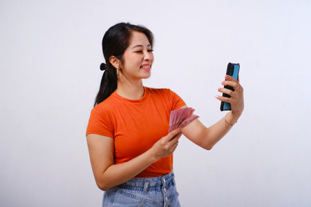 Excited and happy Asian woman wearing casualÂ shirt holding Indonesian money and phone isolated on white backgroundの写真素材