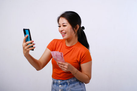 Excited and happy Asian woman wearing casualÂ shirt holding Indonesian money and phone isolated on white backgroundの写真素材
