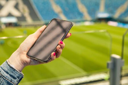 A girl with pink manicure holds a smartphone in her hand against the background of a soccer field,...