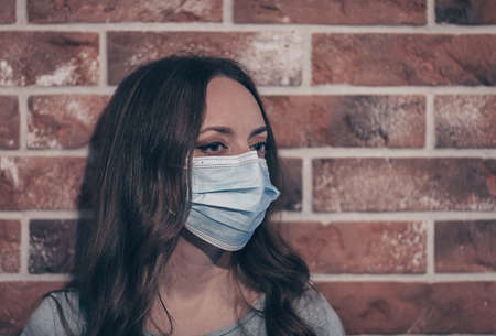 Woman wearing medical face mask to prevent others from corona COVID-19 and SARS cov 2 infection. Portrait of a young woman wearing a blue face mask, looking at camera, on brick background.の写真素材