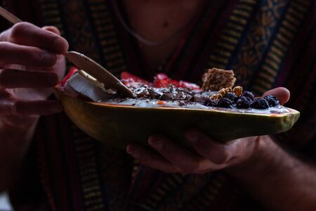 Young man eating healthy breakfast in papaya bowl. Yogurt in papaya bowl with granola, fresh...