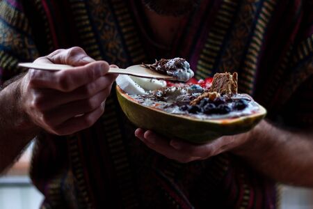 Young man eating healthy breakfast in papaya bowl. Yogurt in papaya bowl with granola, fresh strawberries, blueberries, coconut, chocolate, yogurt and chia seedsの写真素材
