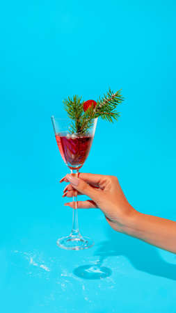 New Year's cocktail party. Cheers! Female hand holding an elegant glass. The glass is filled with a pink cocktail and it's decorated with pine branches and Christmas red and gray glitter balls.の写真素材
