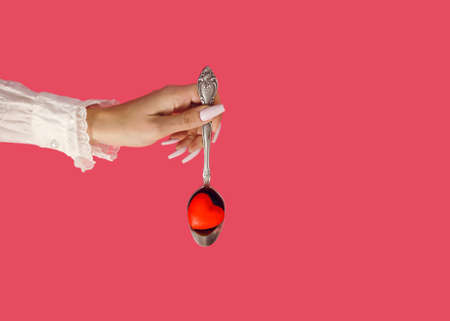 A woman's hand gives a vintage spoon with floral decoration. Love juice and a red heart as symbol of growing love. Modern and creative Valentine's Day flat lay concept. Pink paradise background.の写真素材